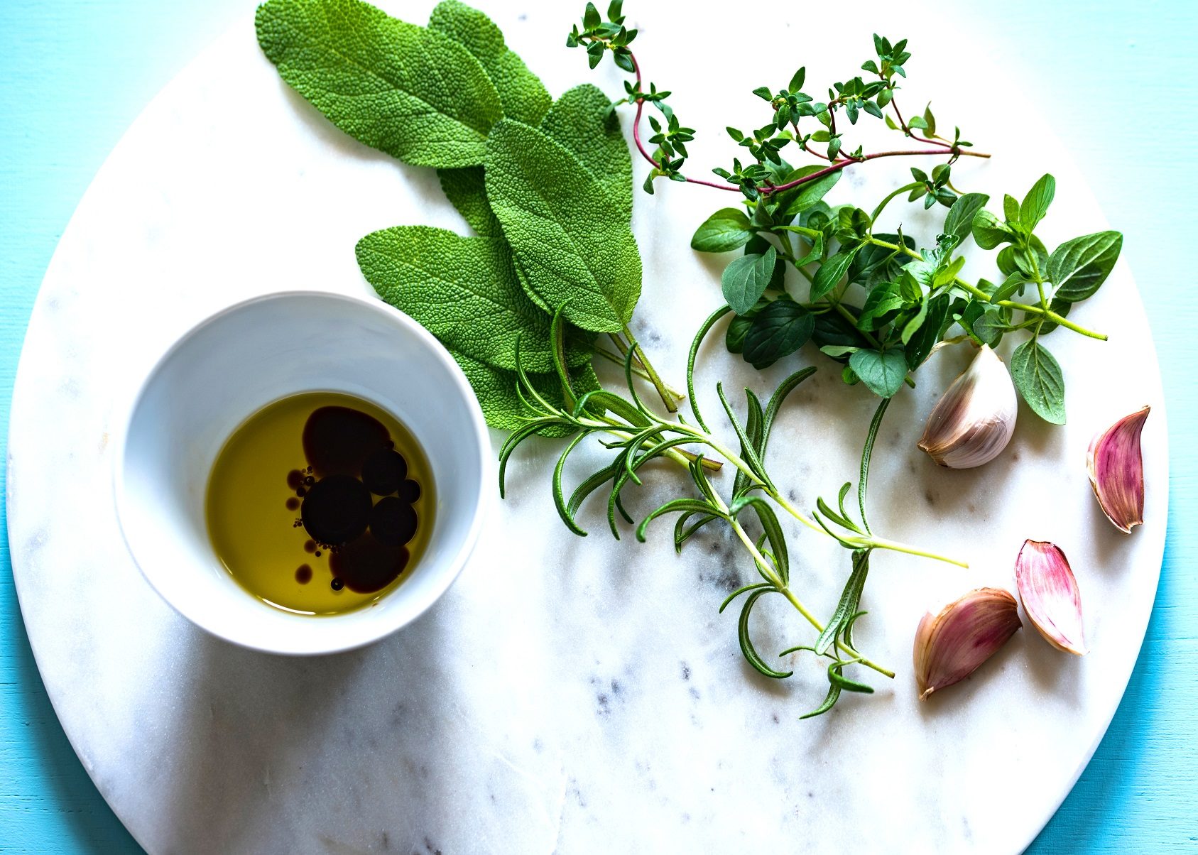 A delicious spread of fresh garlic, sage, rosemary, thyme and oregano on a white marble platter, with a small bowl of balsamic vinegar and olive oil. A delicious no heat options for a high quality and fine oil.